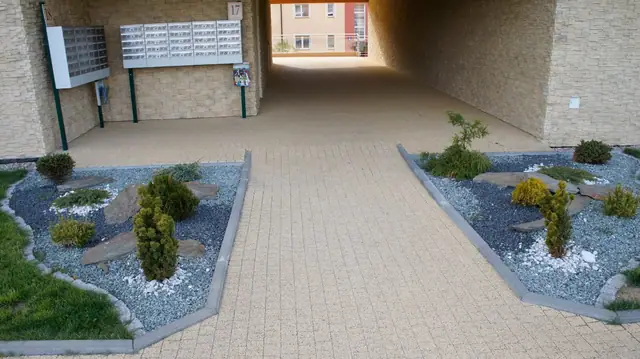 Wybrukowany podjazd prowadzący do przejścia bramowego w budynku, skalniaki po bokach