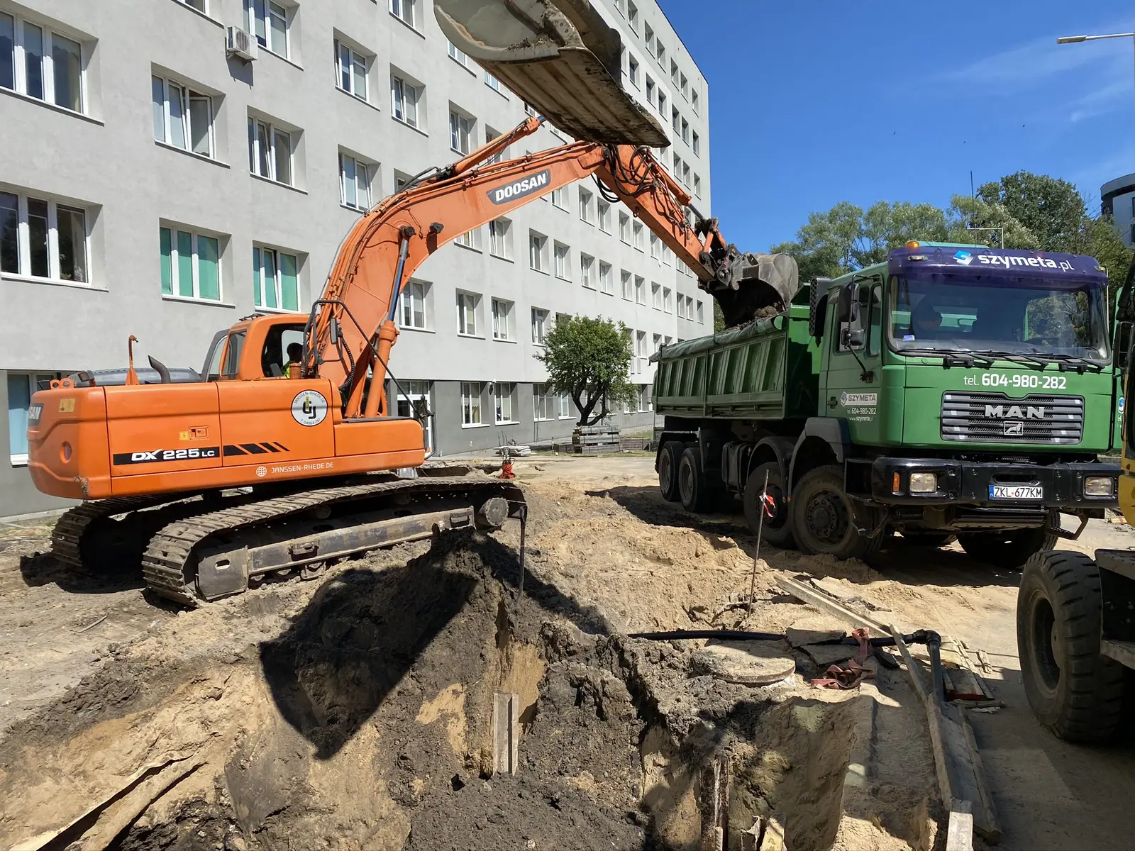 Koparka Doosan ładuje ziemię z wykopu na samochód ciężarowy przy budynku szpitala