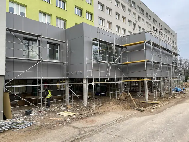 Pawilon szpitala w rusztowaniach podczas montażu elewacji, w tle żółto-zielony budynek