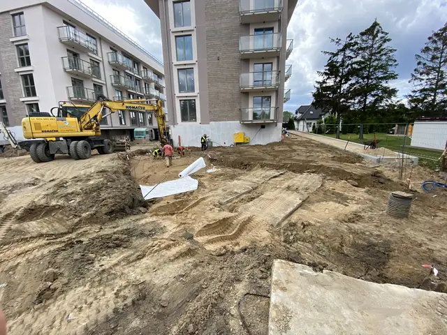 Koparka Komatsu pracuje przy wykopie obok wysokiego budynku z balkonami