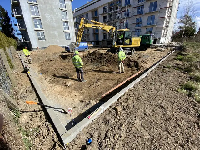 Robotnicy układają krawężnik przy wykopie, w tle koparka Komatsu na placu budowy
