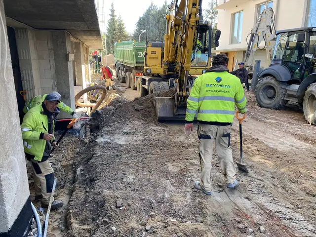 Robotnik firmy Szymeta kopie ręcznie przy elewacji, w tle koparka i ciężarówka z materiałem