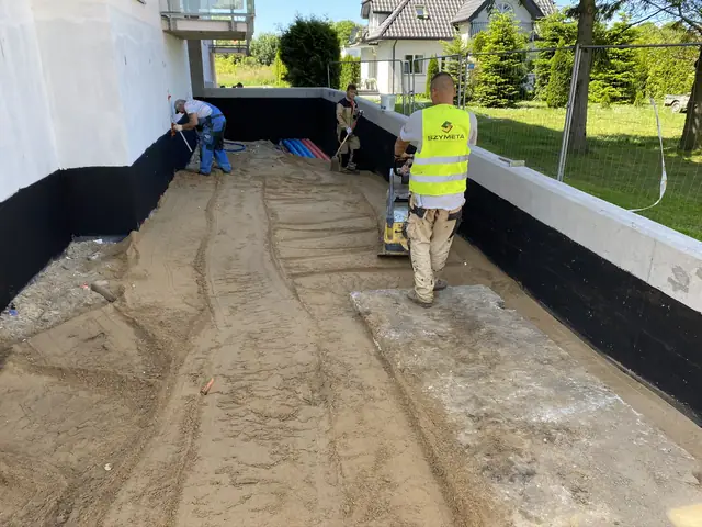 Robotnicy zagęszczają piasek na tarasie przy czarnej hydroizolacji muru oporowego