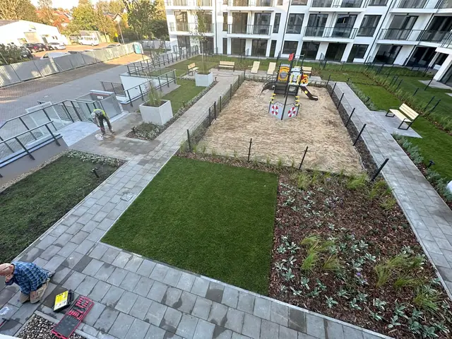 Widok z góry na ogrodzony plac zabaw z piaskownicą i trawnikiem między budynkami osiedla