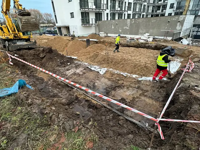 Zabezpieczony taśmą wykop fundamentowy, robotnicy przy pracach ziemnych między budynkami