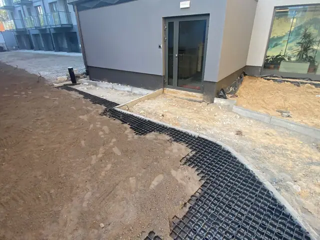 Czarna krata trawnikowa układana przy wejściu do budynku obok pasa piasku