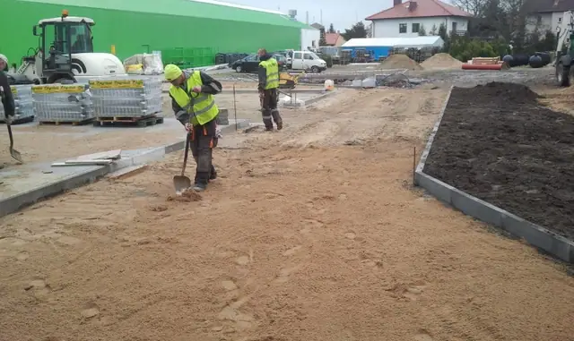 Dwóch robotników przygotowuje podsypkę piaskową wzdłuż krawężnika przed nową halą