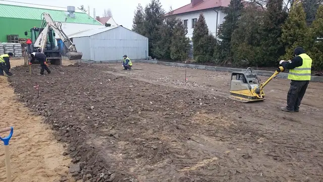 Pracownik zagęszcza wyrównane podłoże zagęszczarką w obecności koparki